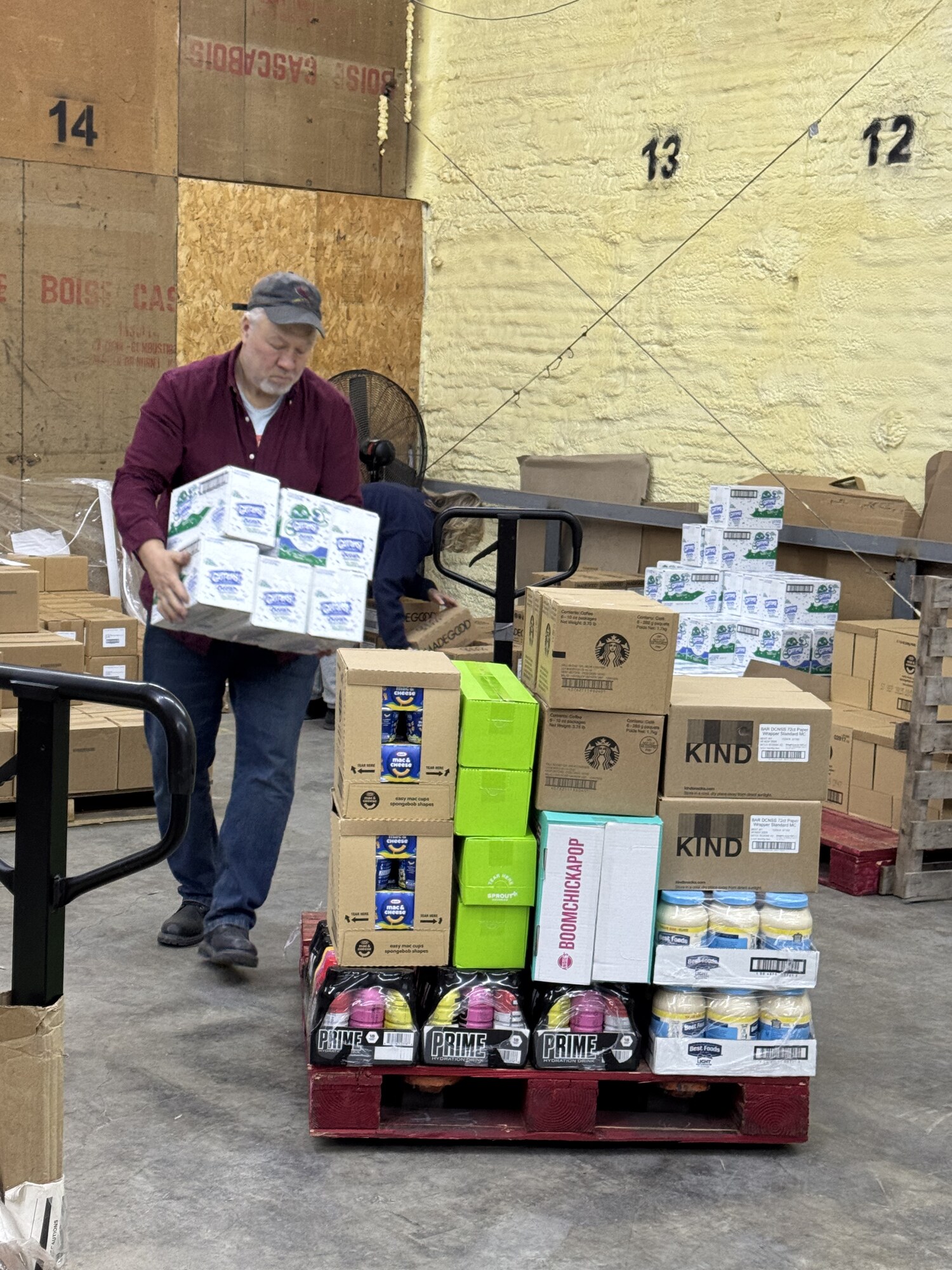 A volunteer carries a case of cheese to a pallet of mac and cheese, snacks, and beverages being assembled inside the warehouse for the next distribution.