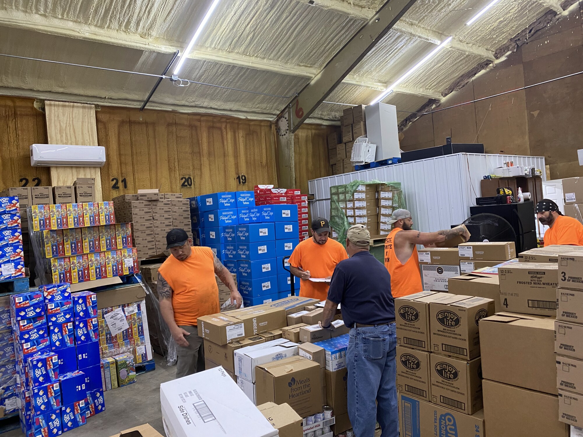 Volunteers in safety orange shirts sort and check pallets of cereal, crackers, and snacks inside the warehouse as they prepare orders for partner agencies.