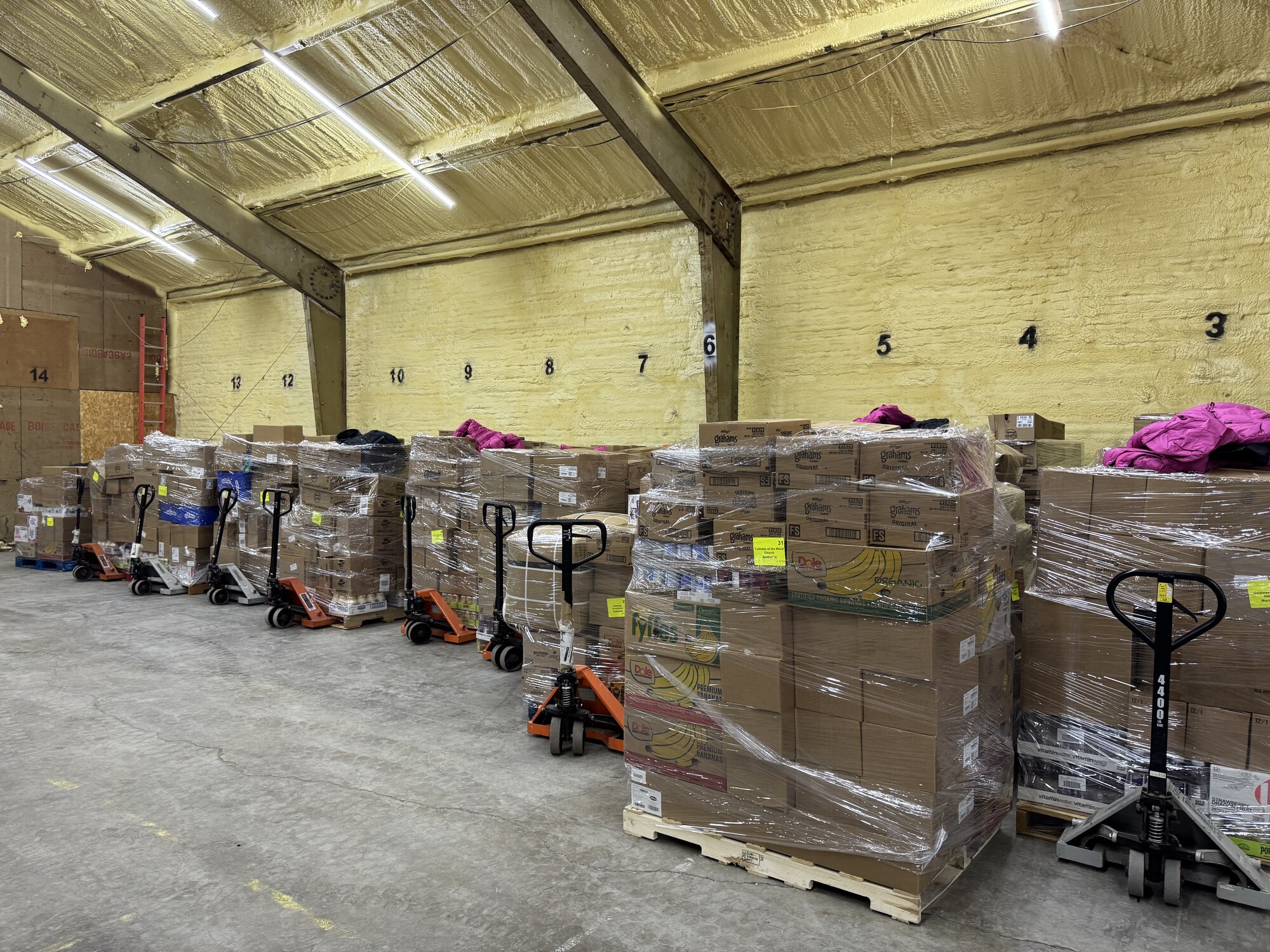 A long row of shrink-wrapped pallets — bananas, graham crackers, vitamin water, and cold-weather coats — staged and ready to be loaded for distribution.