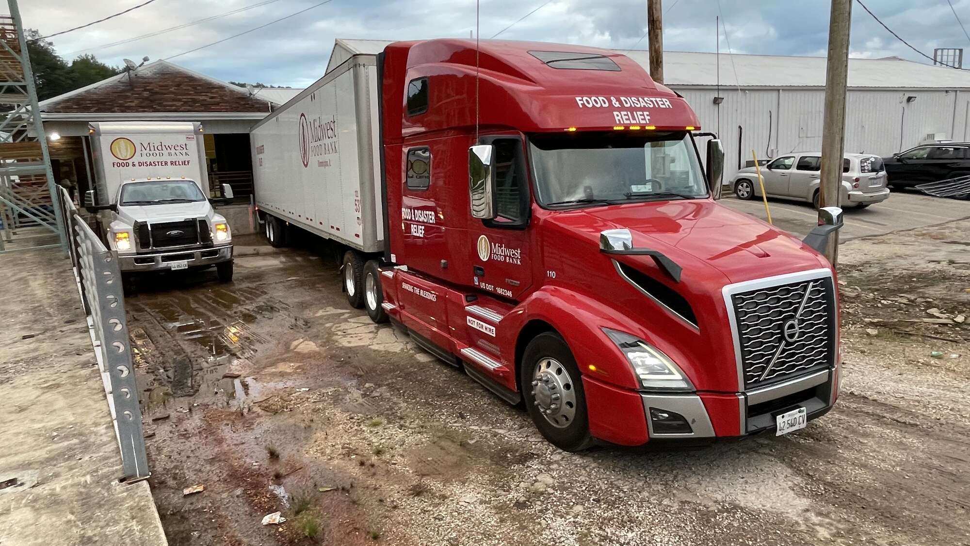 A Midwest Food Bank semi-truck and box truck arrive at the warehouse loading dock to deliver food and disaster relief supplies.
