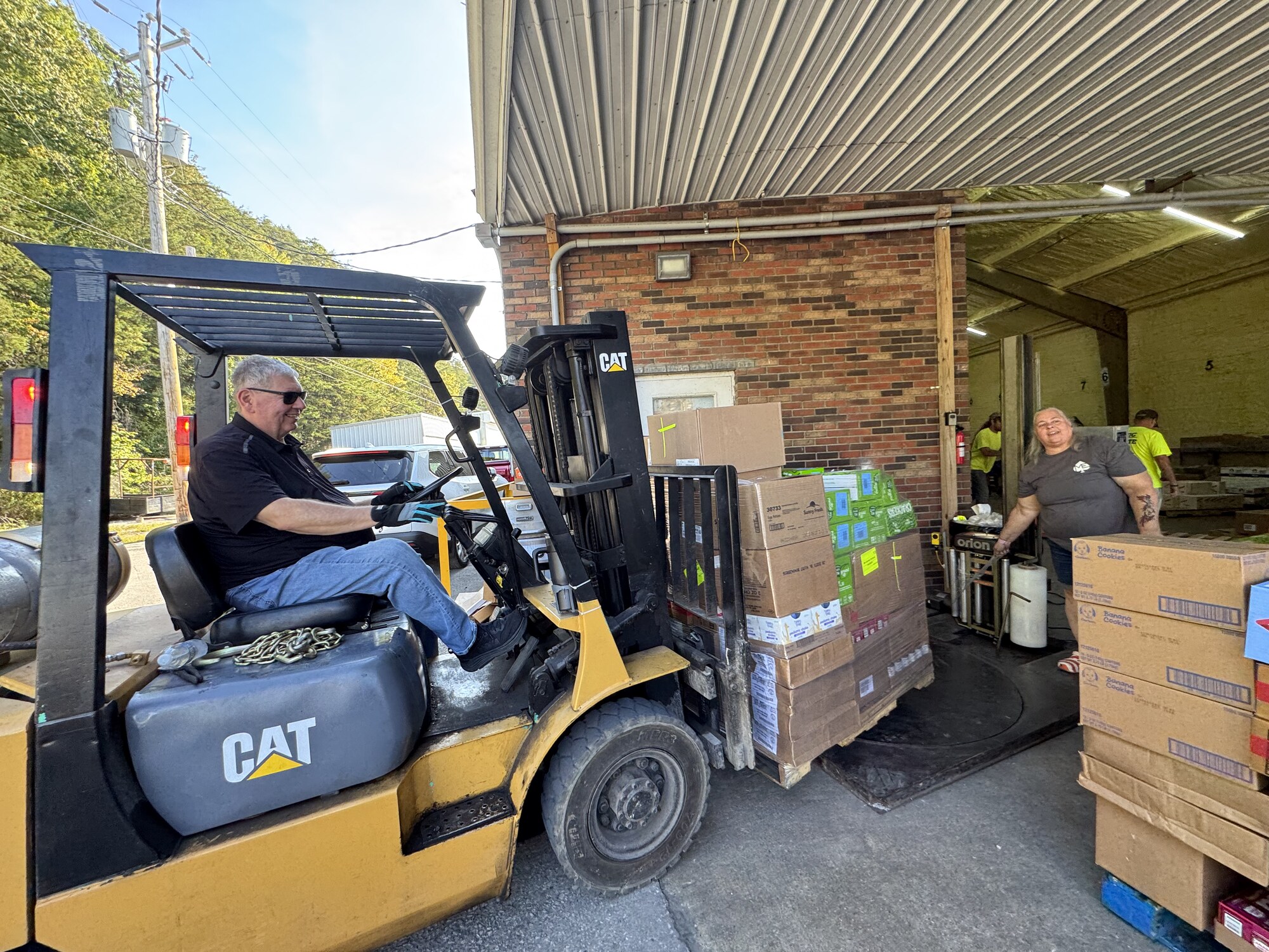 A volunteer drives a forklift carrying a pallet of fresh produce and pantry items out of the loading bay as another volunteer guides it from the dock.