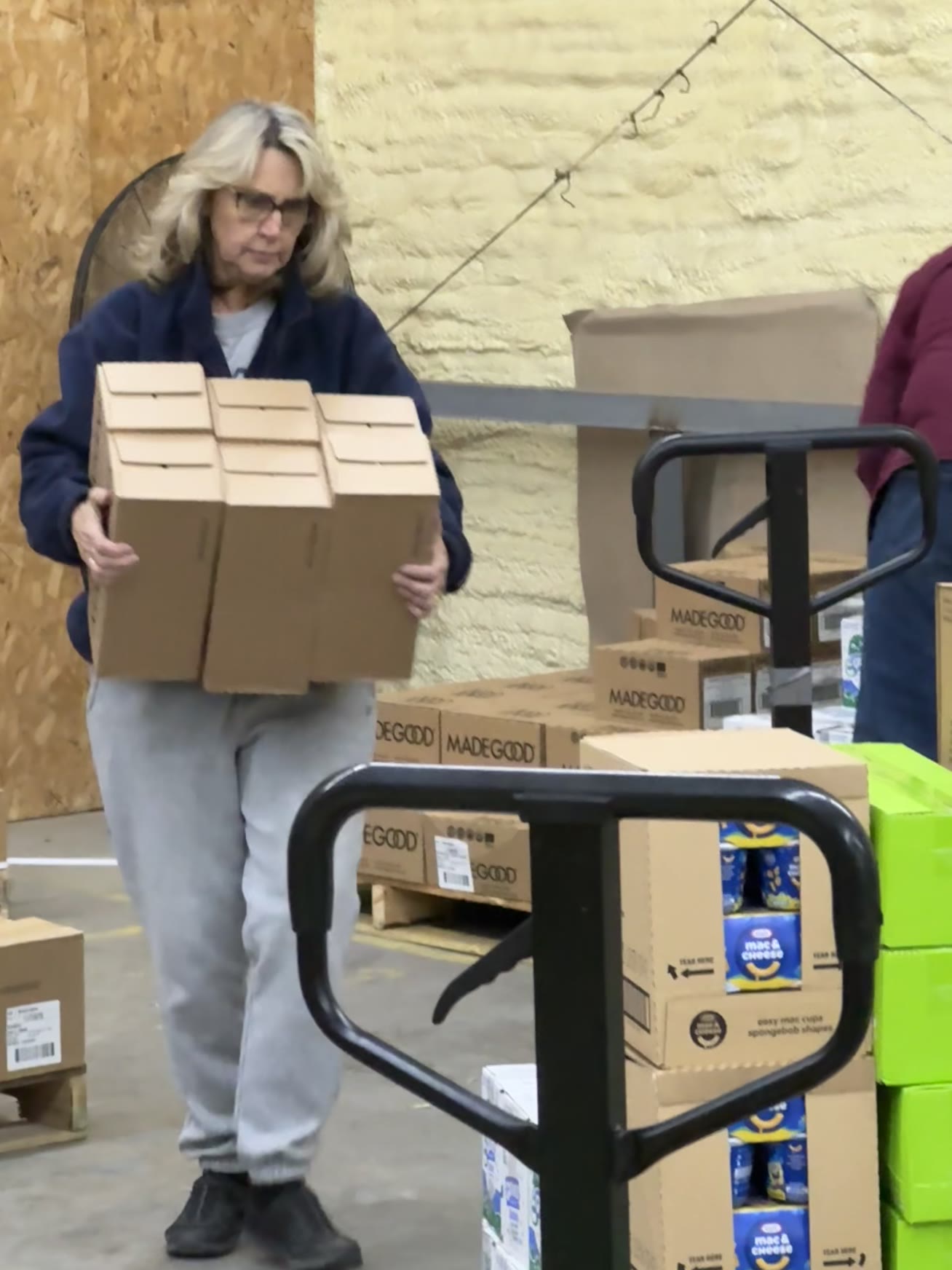 A volunteer carries a stack of empty boxes through the warehouse, preparing to fill them with food for distribution.