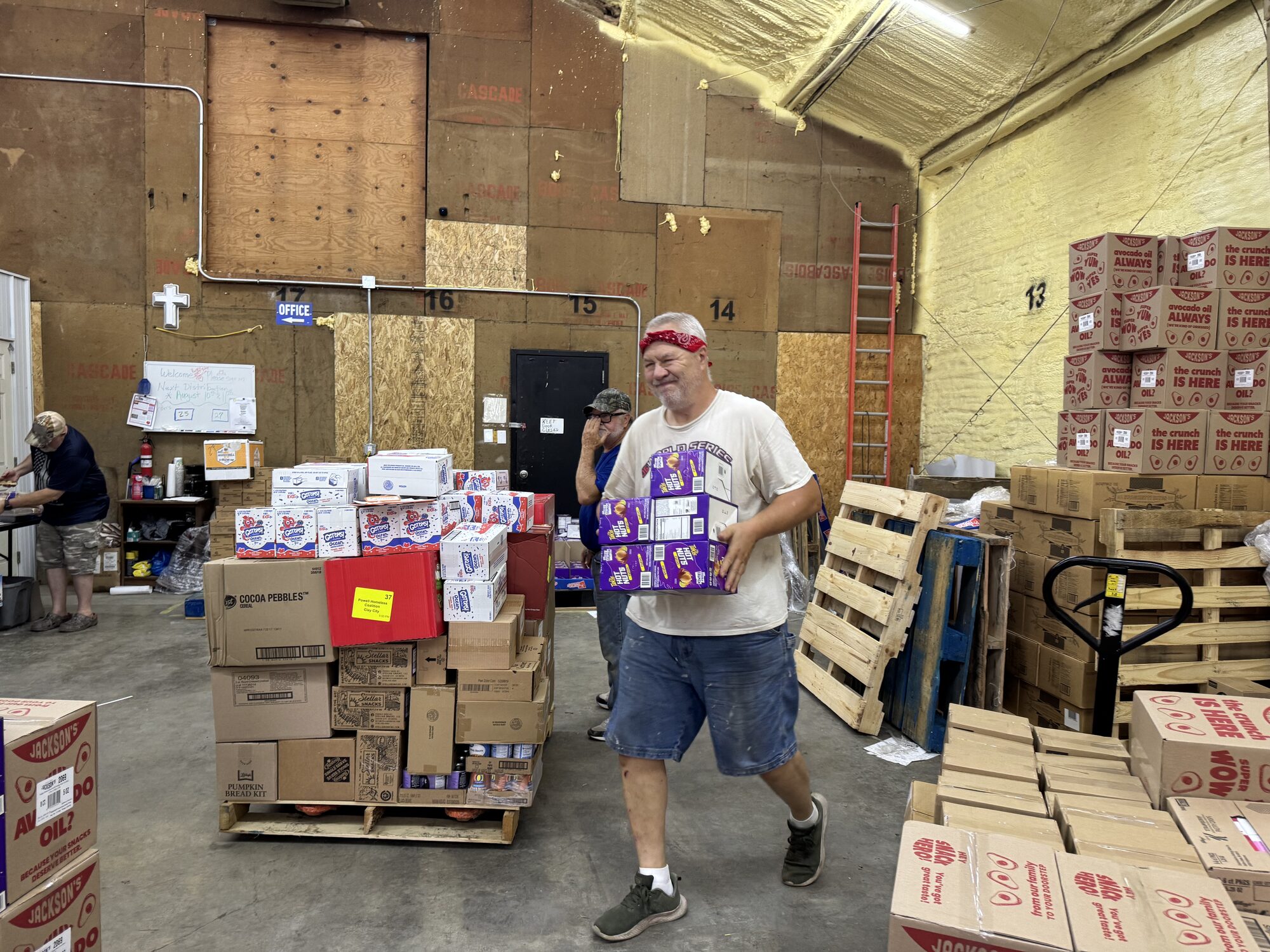 A volunteer carries a case of food across the warehouse floor while others load pallets bound for families in need.